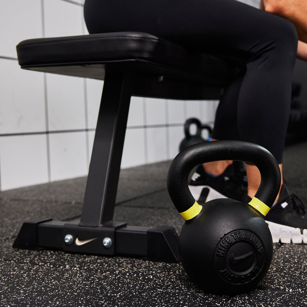 Female athlete sitting on the Nike Flat Weight Bench next to two Nike Kettlebells